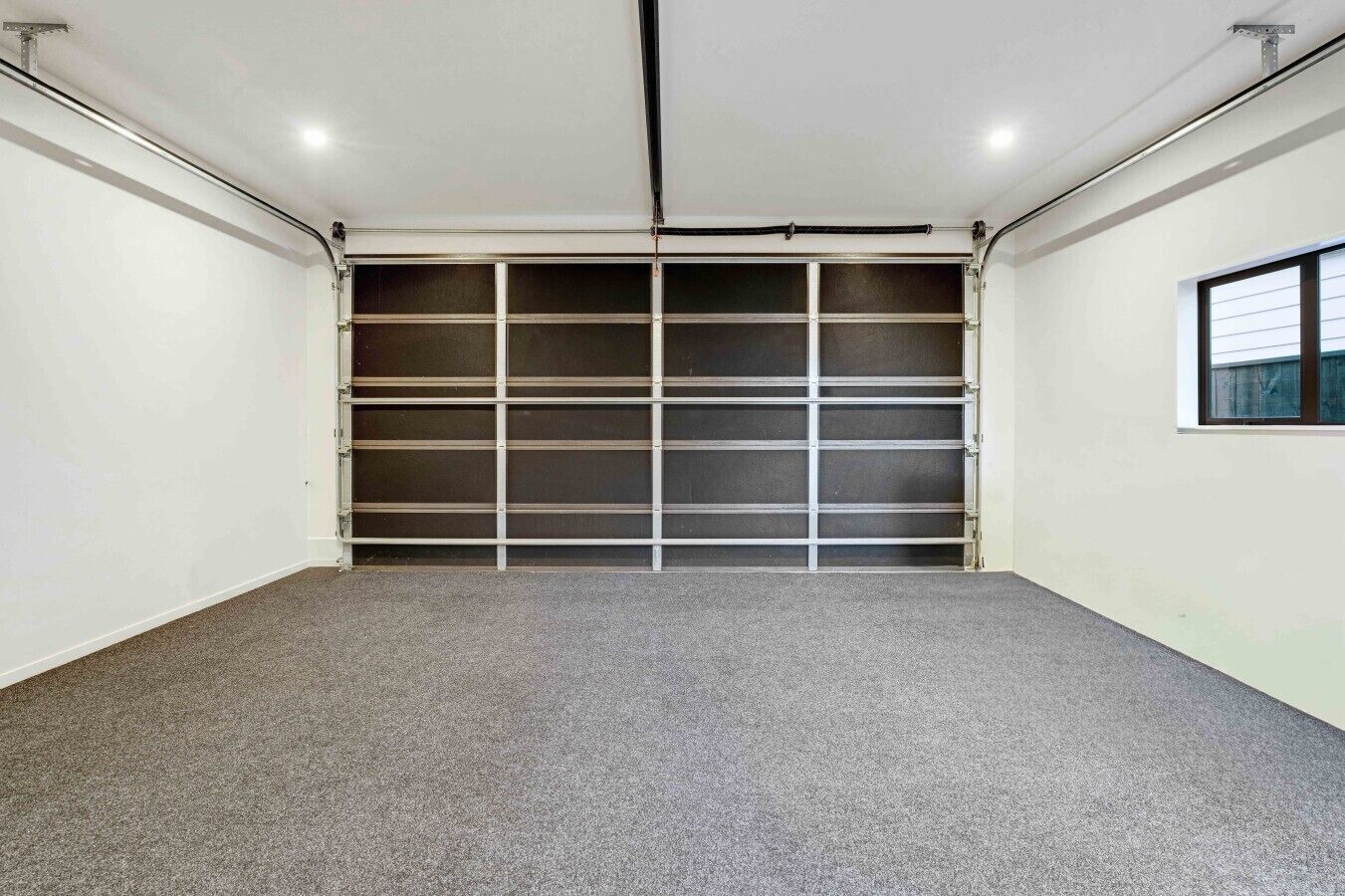 A clean, empty garage with a grey carpet, white walls, and a metal garage door.
