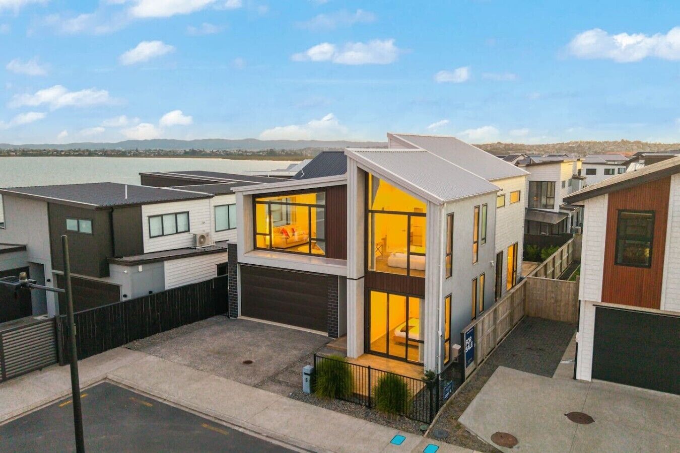 Ein modernes, zweistöckiges Haus mit sichtbarem Schlafzimmer und Wohnbereich, einer Garage und Blick auf das Wasser.