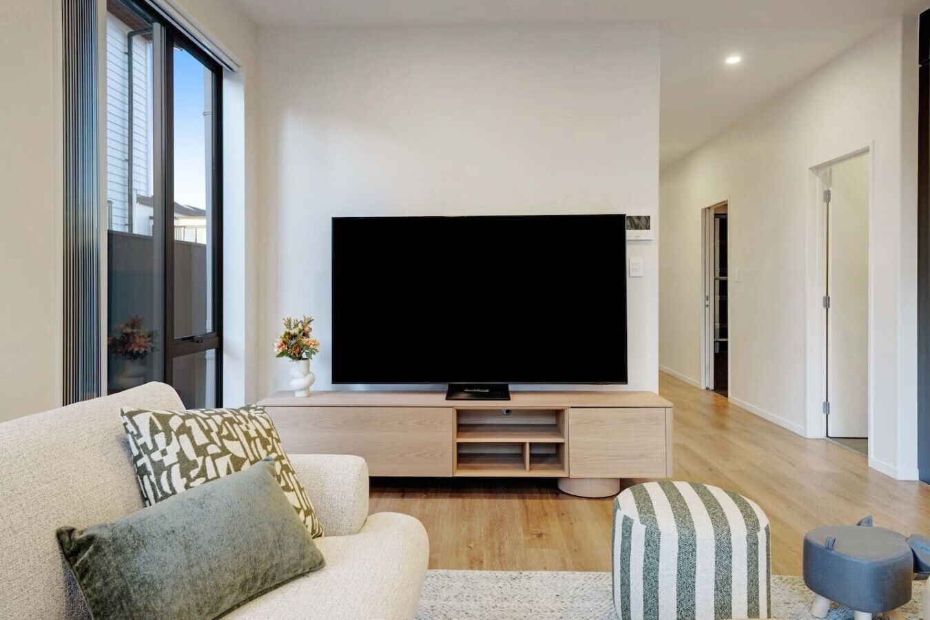 A modern living room with a flat-screen TV on a wooden console, a sofa with patterned cushions, and a small ottoman.