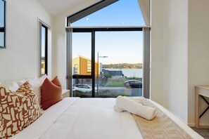 A bedroom with a large bed, a view of a lake, and a modern building in the background. - Hobsonville Family 5BR Sanctuary with Waterviews (Auckland West)