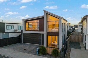 Un immeuble résidentiel moderne à deux étages, doté d’un garage, de grandes fenêtres et d’un intérieur visible, où l’on aperçoit un lit et un canapé.