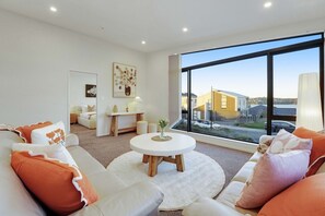Un salon moderne avec une table ronde blanche, des canapés blancs agrémentés de coussins orange, une grande fenêtre donnant sur des immeubles et des voitures, et une décoration minimaliste.
