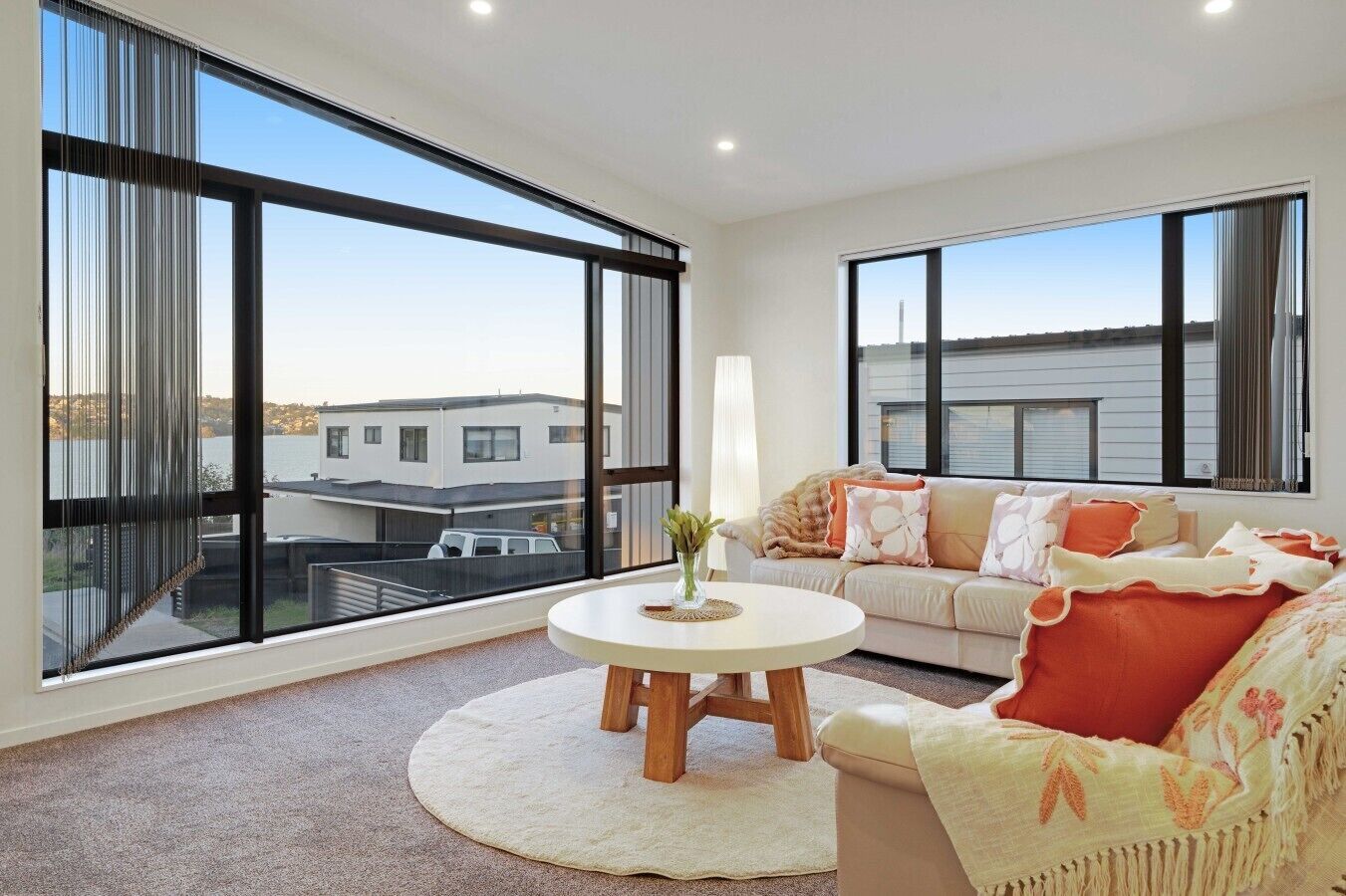 A modern living room with large windows, a round coffee table, a sofa with decorative pillows, and a view of buildings outside.