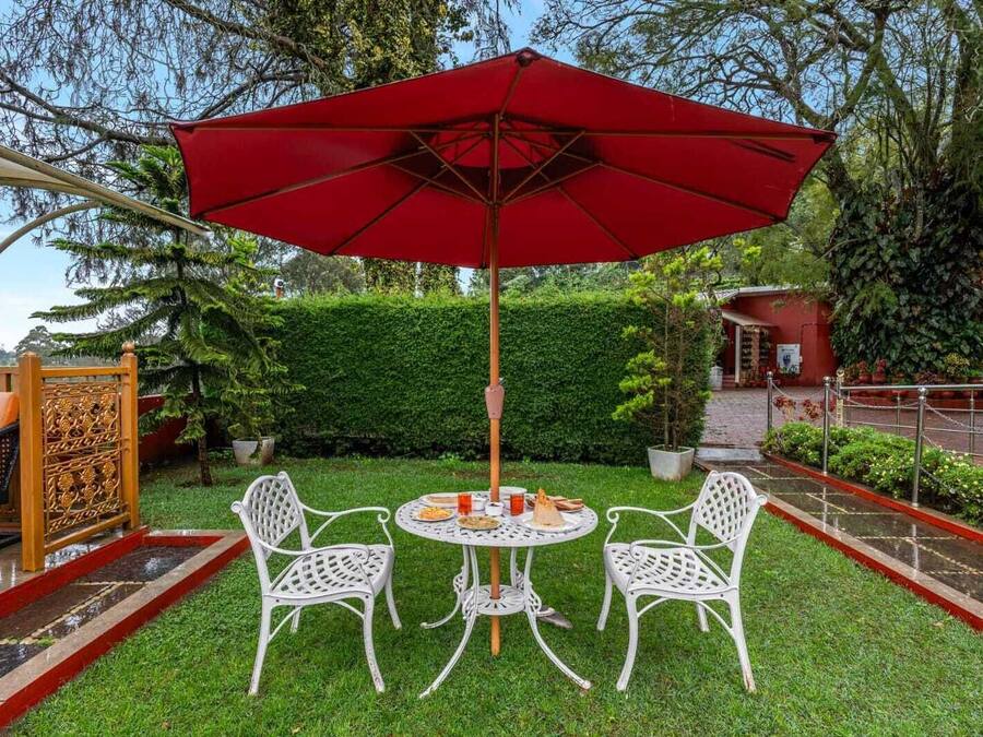 A round table with four white chairs and a red umbrella in a garden setting.