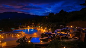Vista nocturna de un resort con piscina, toboganes acuáticos y montañas al fondo.
