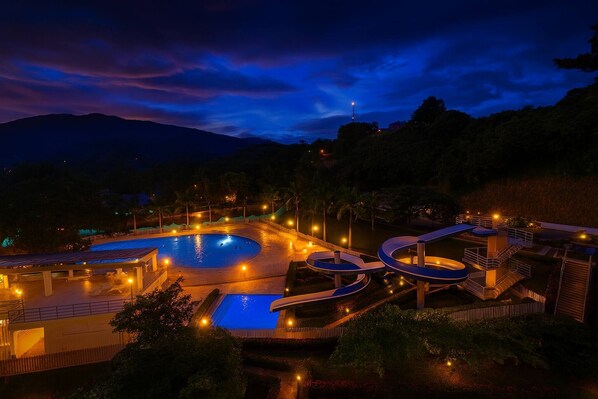 A nighttime view of a resort with a swimming pool, water slides, and a mountain backdrop.
