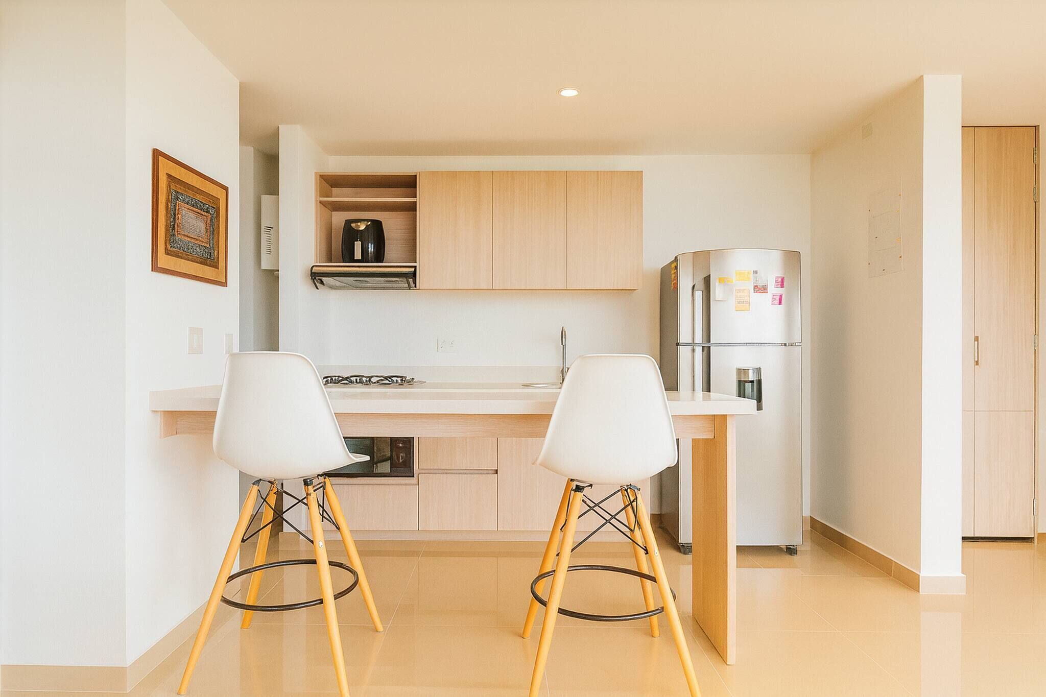 Una cocina moderna con dos taburetes blancos, un refrigerador de acero inoxidable y armarios de madera clara.