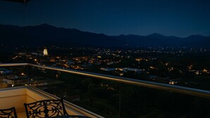 A balcony with a table and chairs, a view of a cityscape at night, and mountains in the distance.