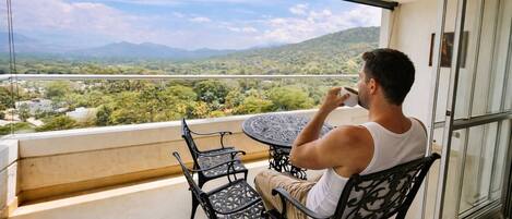 Una persona sentada en una mesa en un balcón con vista a un paisaje verde y montañas.