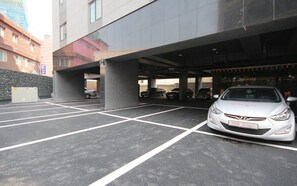 A parking garage with multiple parking spaces, a white car parked in one, and a building with a modern facade in the background.