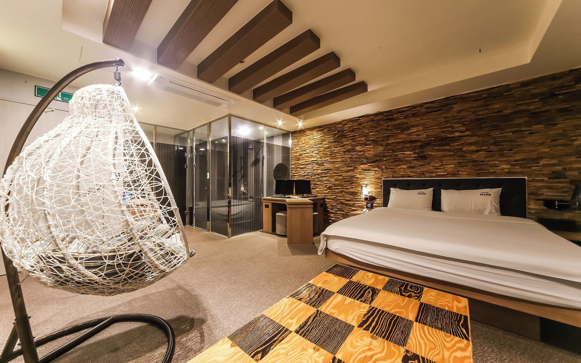 A modern hotel room with a bed, a desk, a hanging chair, and a stone wall.