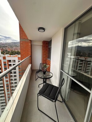Ein Balkon mit Glasdoor, einem runden Tisch, zwei Stühlen, einem Blumentopf und Blick auf eine Stadtlandschaft.