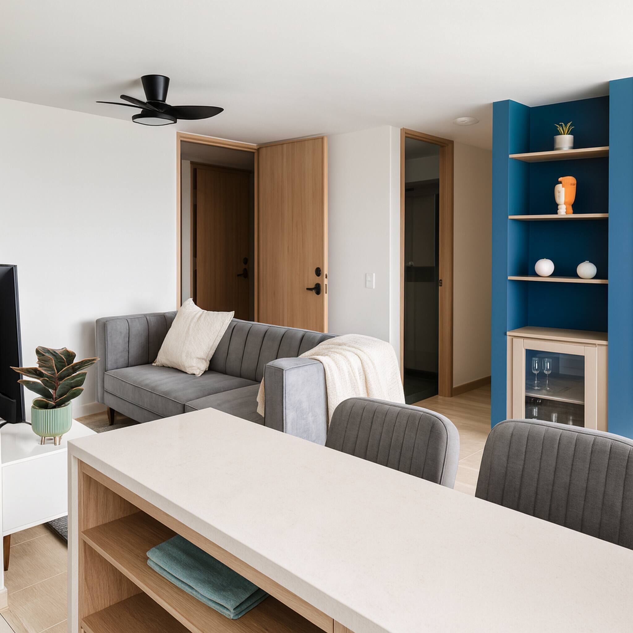 A modern living room with a sofa, a dining table, and a TV. There is a ceiling fan and a bookshelf with decorative items.