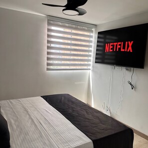 Ein Schlafzimmer mit einem Bett, einem Deckenventilator, einem Fenster mit Jalousien und einem an der Wand befestigten Fernseher, der das Netflix-Logo zeigt.
