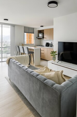 A modern living room with a grey sofa, a flat-screen TV, and a dining area with chairs and a kitchen in the background. - Epic View 25 Pablo Tobón Cardiovascular (Medellín)