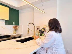 A person in a robe pouring a drink from a bottle into a glass at a kitchen counter with a modern faucet and marble countertop. - Green Lux Garden - 2BR 1Bath - Free Parking (Calgary)