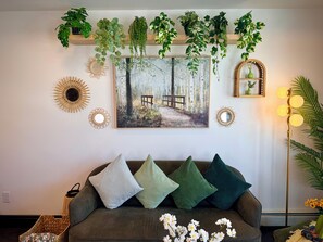 Uma sala de estar com sofá, almofadas decorativas, obras de arte nas paredes, plantas penduradas e um abajur.