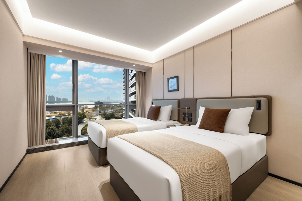 Habitación de hotel con dos camas, un ventanal con vistas a la ciudad y un televisor de pantalla plana en la pared.