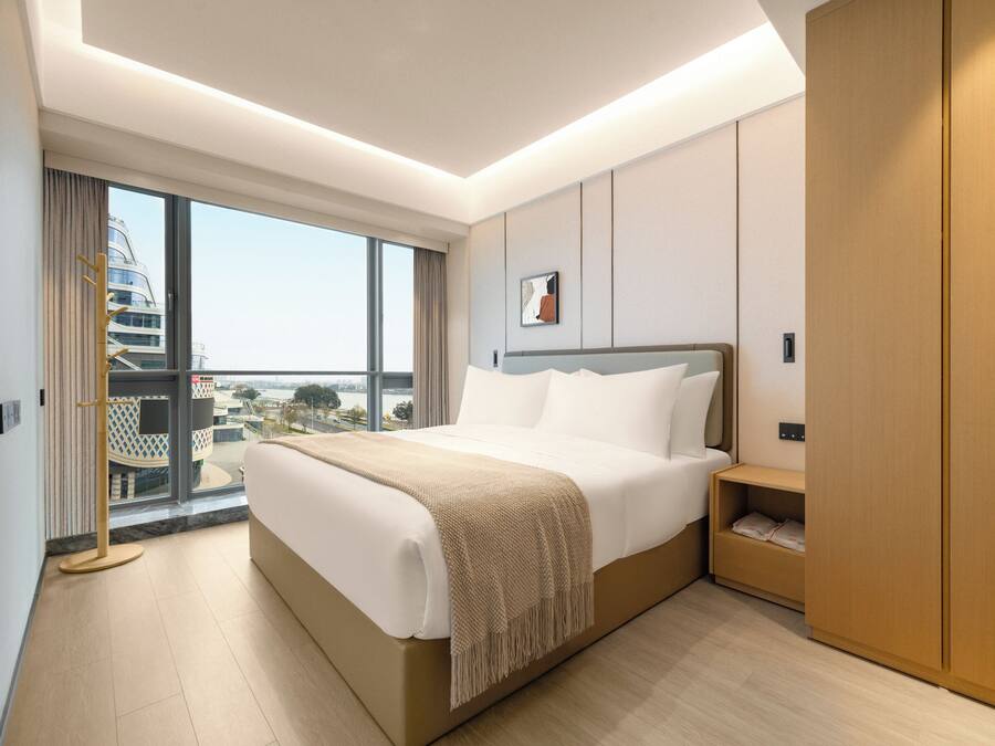 A modern hotel room with a large bed, a wooden wardrobe, and a view of a cityscape through a large window.