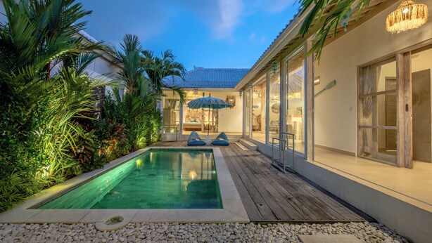 Un área de piscina con terraza de madera, una sala con paredes de cristal y plantas tropicales.