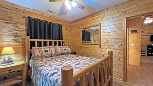 Une chambre dans une cabane en bois, avec un lit, une table de chevet, une lampe, un ventilateur de plafond, une fenêtre avec des rideaux, un miroir et une porte.