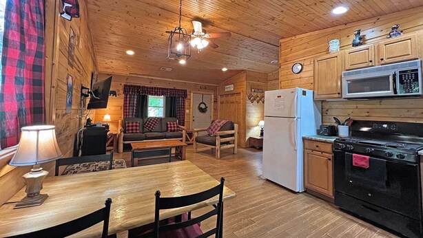 Un intérieur de chalet chaleureux avec une table à manger en bois, des chaises, un canapé, une cuisine équipée d’une cuisinière et d’un four à micro-ondes, et un ventilateur de plafond.