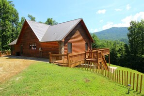 A log cabin with a wooden deck and a metal roof, surrounded by a fenced yard and lush greenery. - Glory on the Mountain (Bryson City)