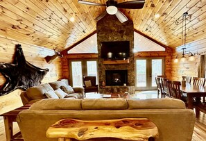 A log cabin interior with a stone fireplace, leather sofa, and a dining area. - Glory on the Mountain (Bryson City)