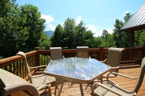A deck with a glass-top table and chairs, surrounded by a wooden railing, overlooking a forested area. - Glory on the Mountain (Bryson City)