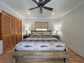 Un dormitorio con cama, cabecero de madera, dos mesitas de noche con lámparas, un ventilador de techo y una ventana con persianas.