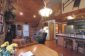 Ein rustikales Interieur mit Holzbalkendecke, einem Essbereich mit Holztisch und -stühlen, einem Wohnzimmer mit einer Couch und einer Küche mit Kühlschrank und Herd.