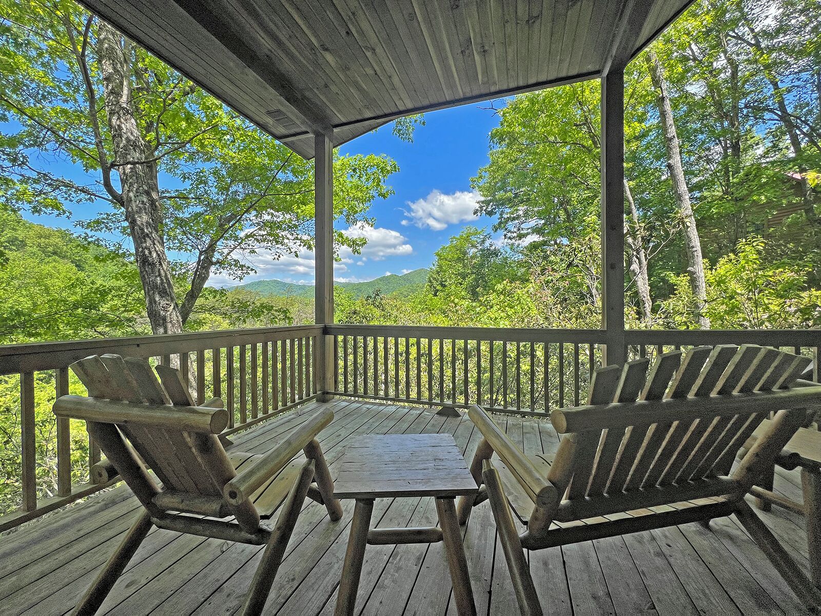 Une terrasse en bois avec deux chaises et une table, donnant sur une zone boisée avec des montagnes à l’horizon.