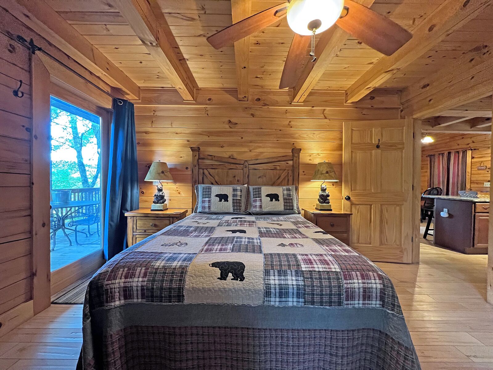 A log cabin bedroom with a bed, two nightstands, a ceiling fan, and a view of the outdoors.