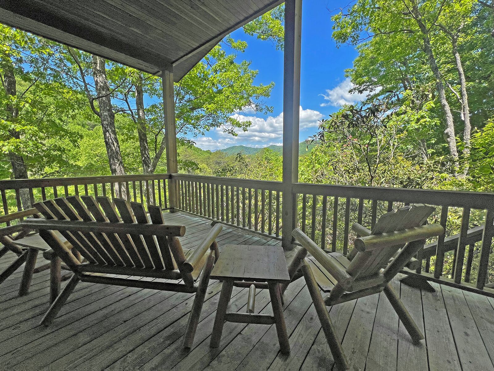 Eine überdachte Holzterrasse mit Adirondack-Stühlen und einem Tisch, umgeben von Bäumen und blauem Himmel.