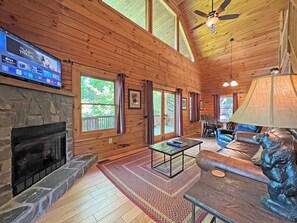 Ein Interieur einer Holzhütte mit Kamin aus Stein, einem an der Wand montierten Fernseher, einem Deckenventilator und einem Essbereich mit Tisch und Stühlen.