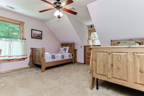 Ein Schlafzimmer mit schrägem Dach, einem hölzernen Bett, einer Holzkommode, einem Fenster mit Vorhängen und einem Deckenventilator.