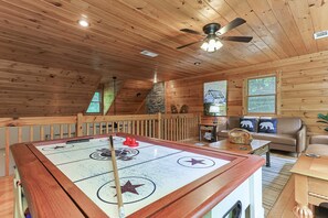 Ein Holzinnenraum mit einem Shuffleboard-Tisch, einem Deckenventilator, einer Couch und einem Fenster mit Blick auf Bäume.