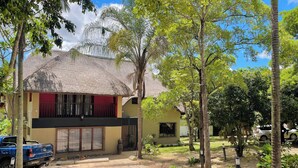 A building with a thatched roof, a paved entrance, and a small garden area with trees and a car parked nearby.