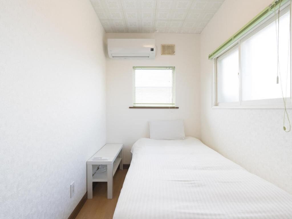Une petite chambre propre, avec un lit, une table de chevet, une fenêtre avec des stores et un climatiseur fixé au mur.