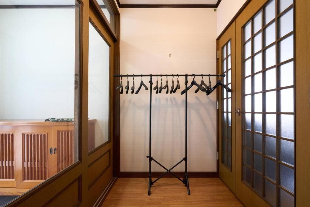 Una habitación con piso de madera, un perchero de metal negro con ropa colgando y un ventanal de vidrio corredizo con paneles esmerilados.