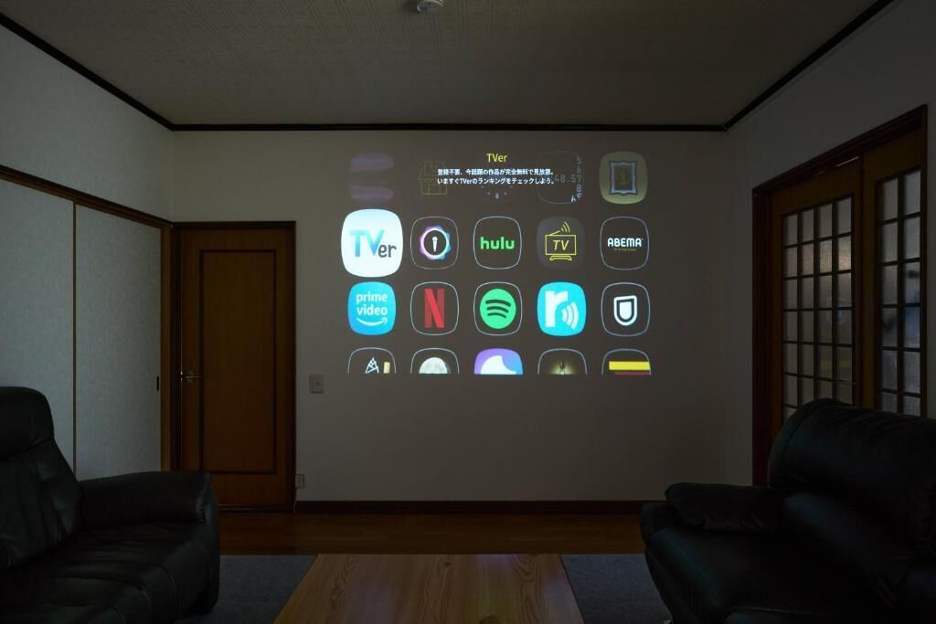 A room with a wall-mounted projection displaying various streaming service icons. There are two black leather sofas and a wooden floor.
