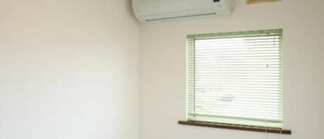 A bedroom with a single bed, a bedside table, a window with blinds, an air conditioning unit, and a wall-mounted vent.