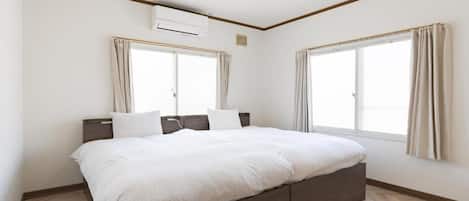 A bedroom with a bed, two windows with curtains, an air conditioning unit, and a herringbone-patterned floor.