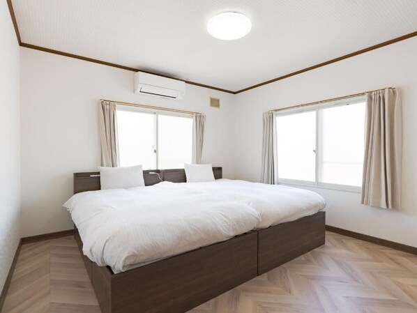 A bedroom with a bed, two windows with curtains, an air conditioning unit, and a herringbone-patterned floor.