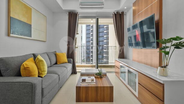 Un soggiorno moderno con un divano grigio, un tavolino in legno, una televisione e un balcone con vista su edifici alti.