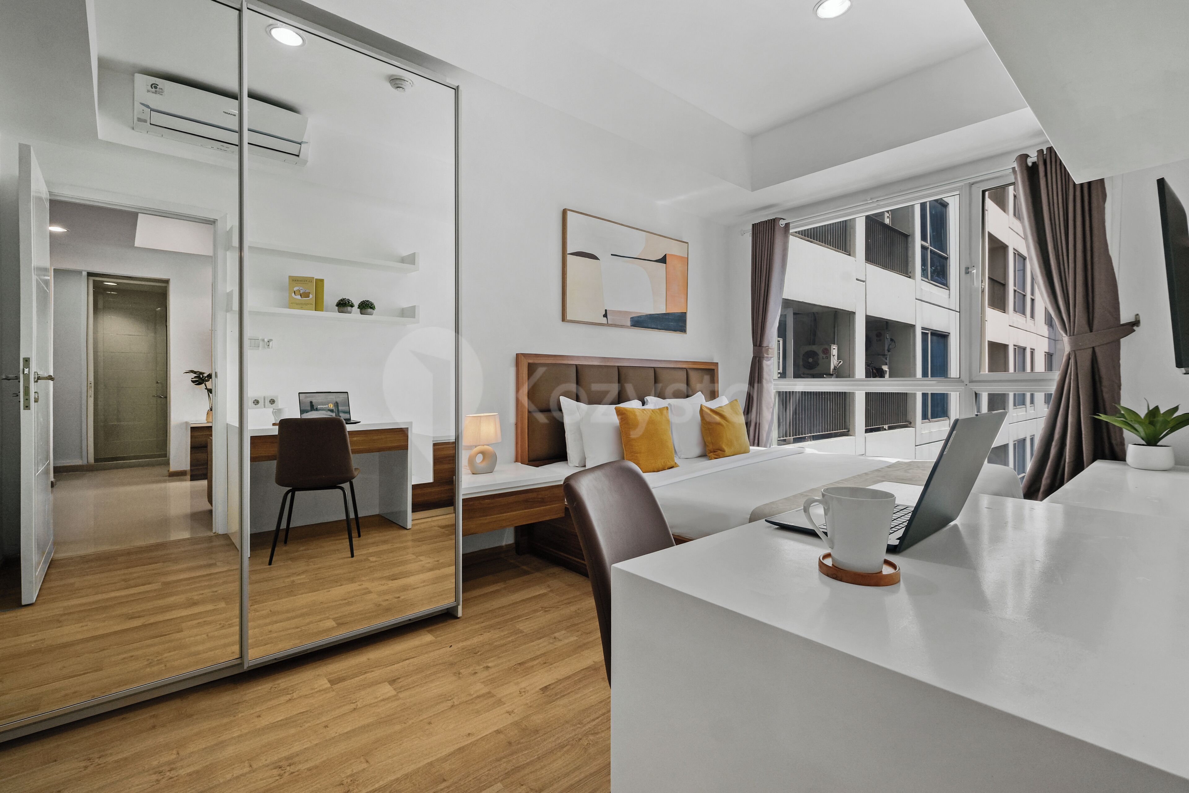 Une chambre d’hôtel moderne avec une cloison en verre, un bureau avec un ordinateur portable, un lit avec des oreillers et une grande fenêtre avec des rideaux.