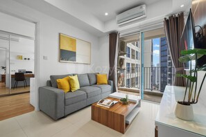 A modern living room with a grey sofa, a wooden coffee table, and a large window with curtains. - Astera by Kozystay - Kuningan (South Jakarta)