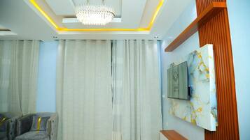 A modern living room with a chandelier, curtains, a TV mounted on a marble wall, and a grey sofa.