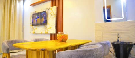 A modern dining area with a wooden table, grey upholstered chairs, a decorative bowl, a wall-mounted TV, and a unique black sink.