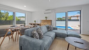 Een moderne woonkamer met een grijze modulaire bank, een eettafel met stoelen, een klein houten kastje en uitzicht op een zwembad door grote ramen.